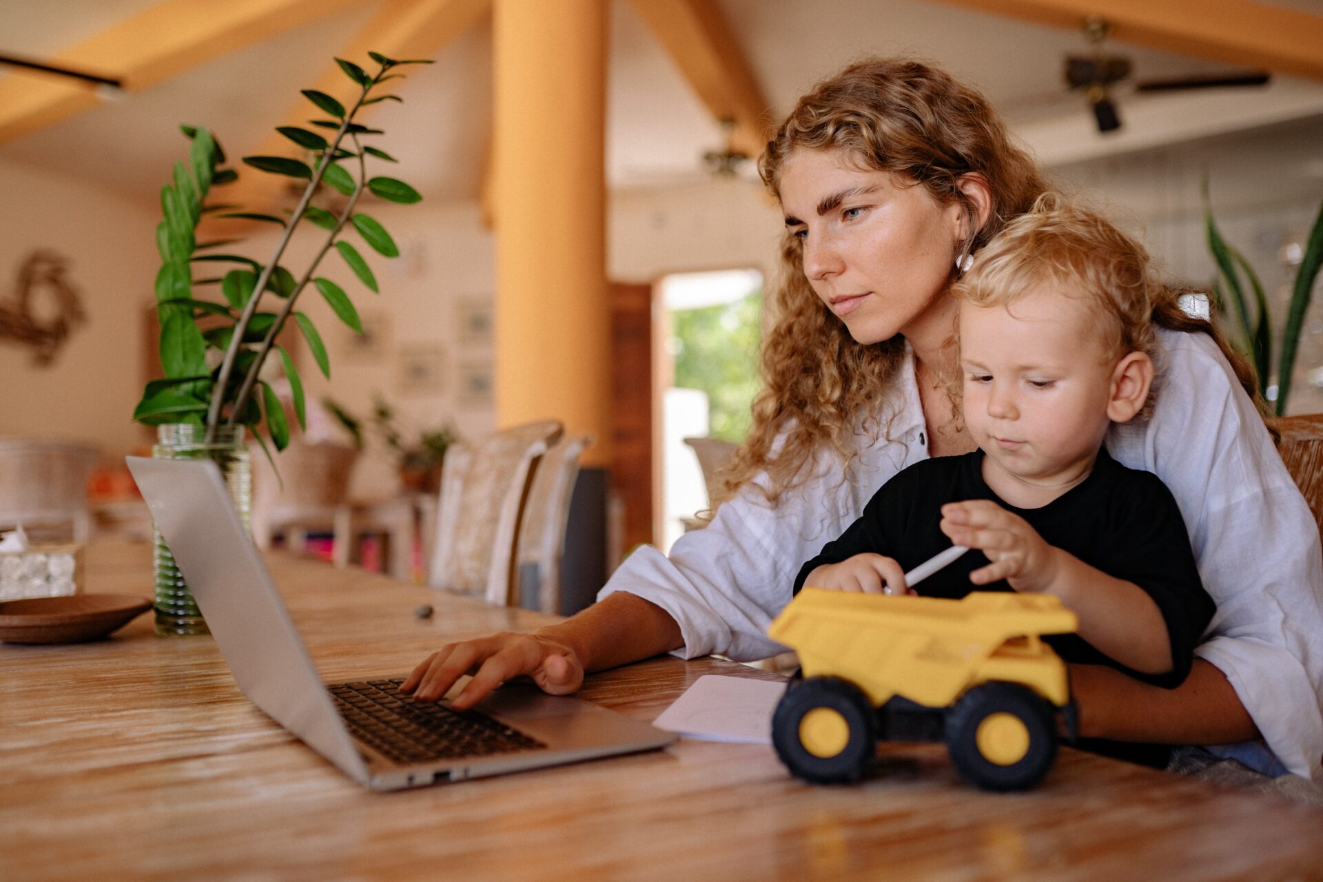 A mother using her laptop whilst holding her son who is playing with a yellow toy truck.