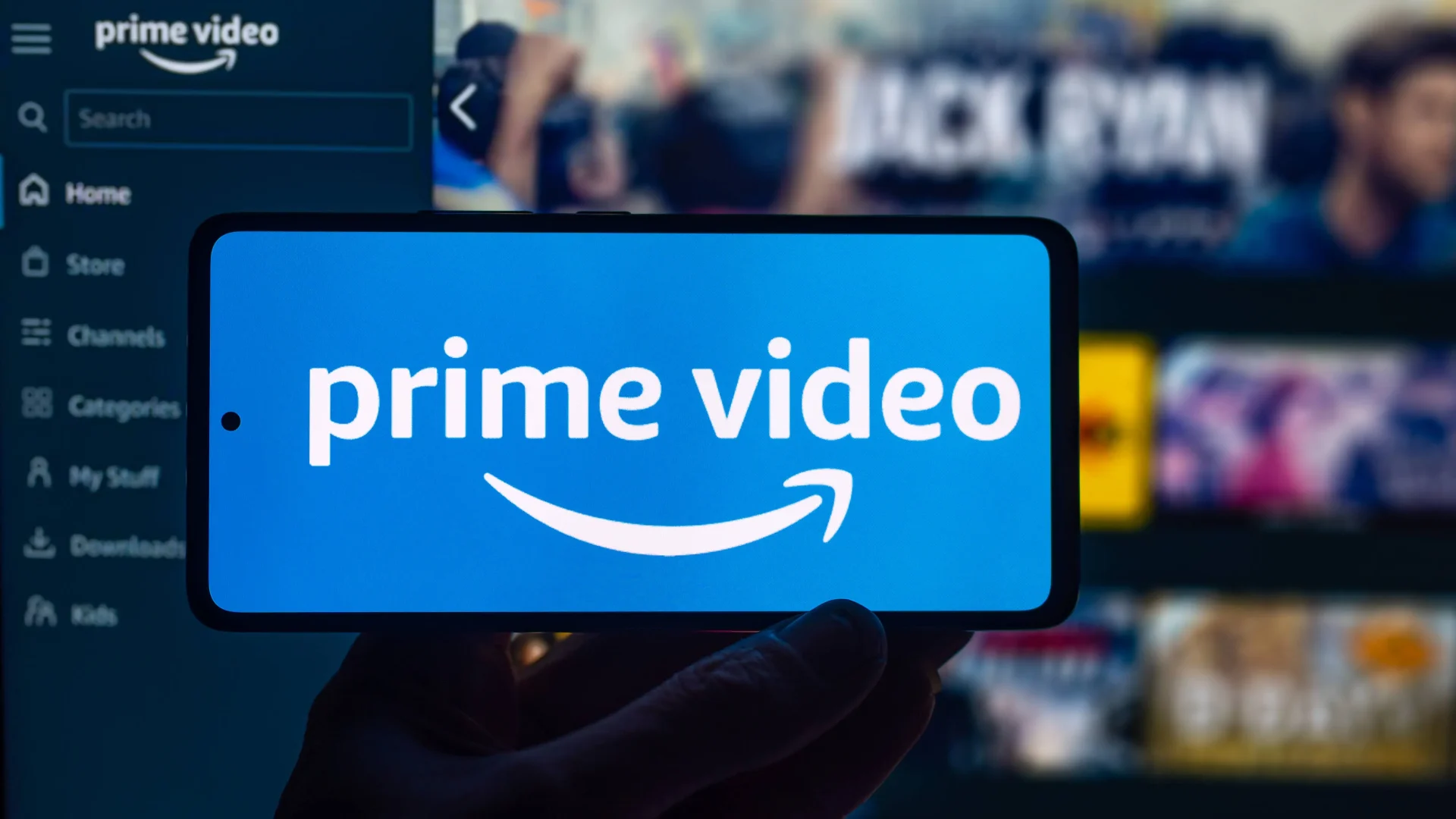 A hand holding up a smartphone screen with the Amazon Prime Video logo, with the Amazon Prime Video application in the background.